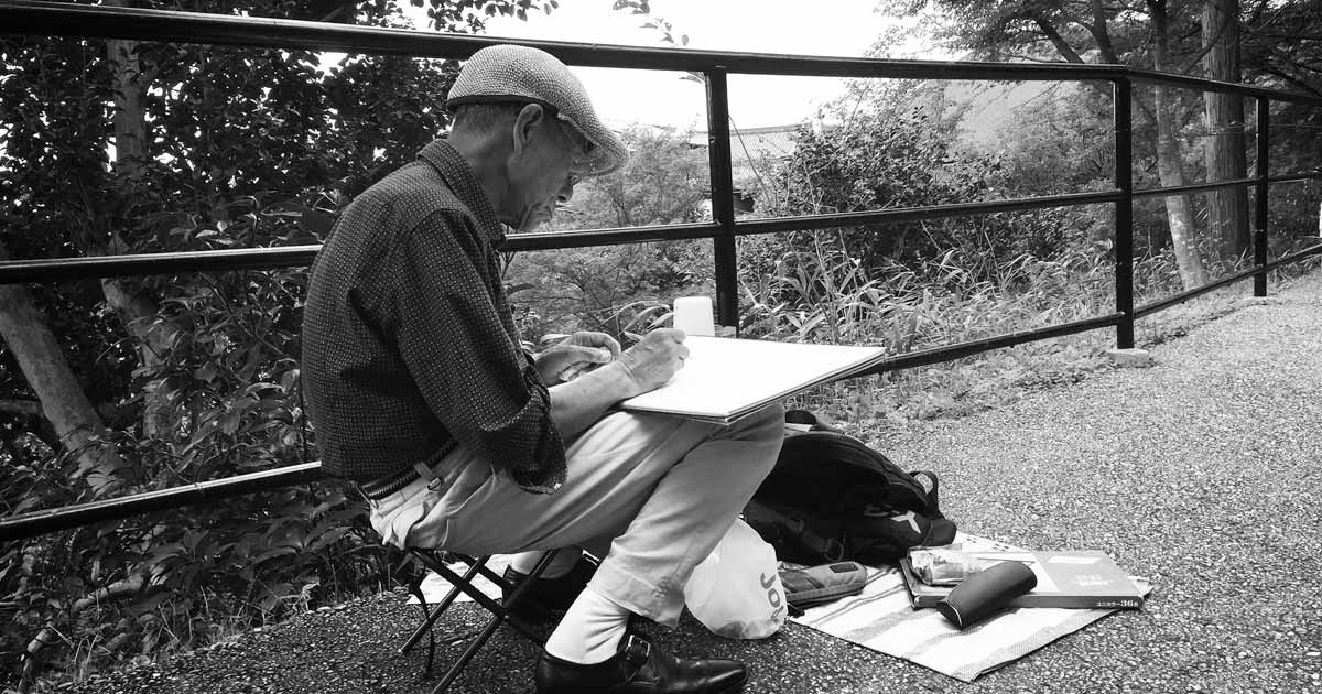 An elderly man sitting in a small seat drawing in a notebook.